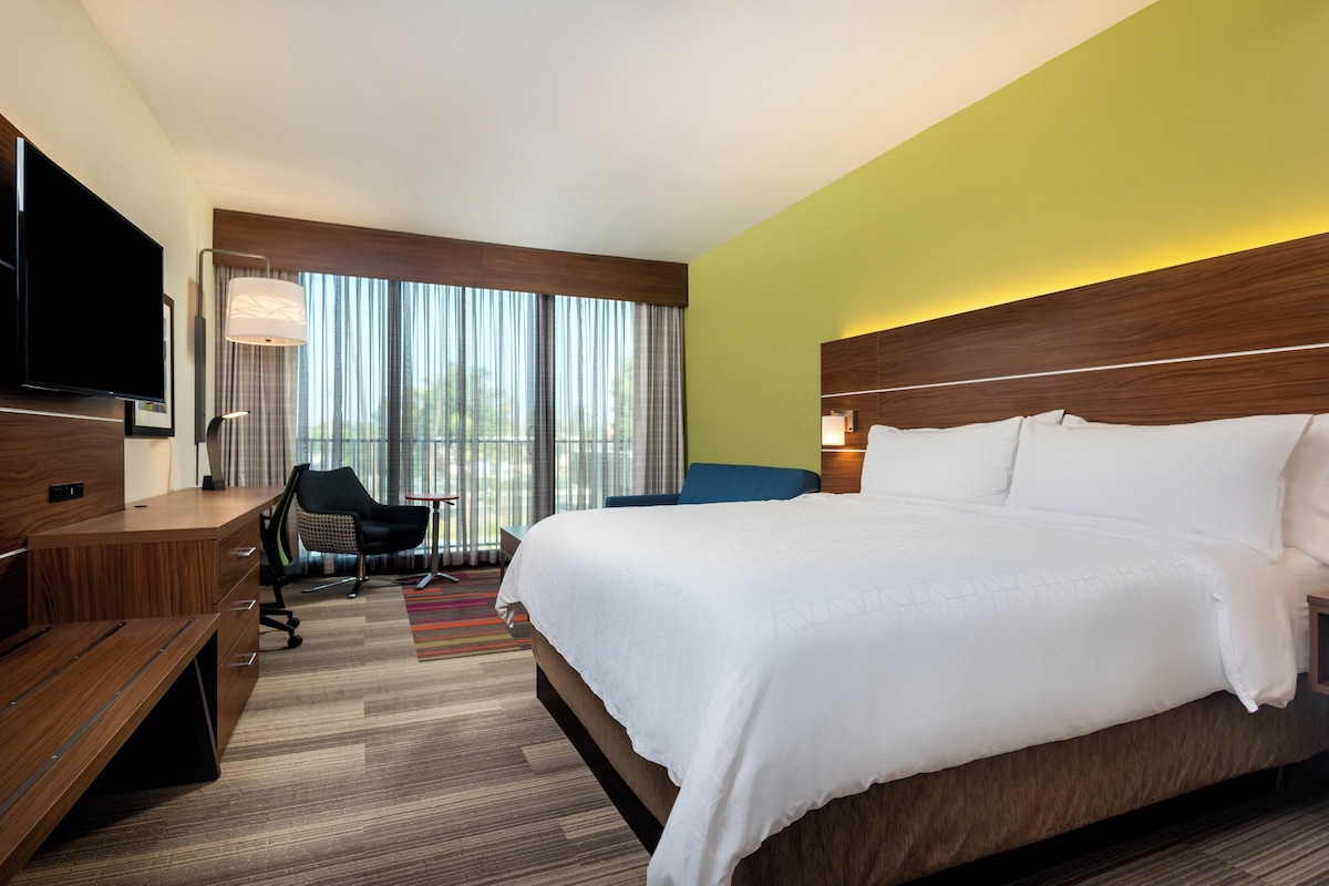 Habitación de hotel con una cama grande, un escritorio con silla, televisión y ventana con cortinas.