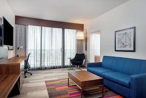 Habitación de hotel moderna con escritorio de madera, sofá azul, alfombra a rayas colorida y un ventanal con cortinas.