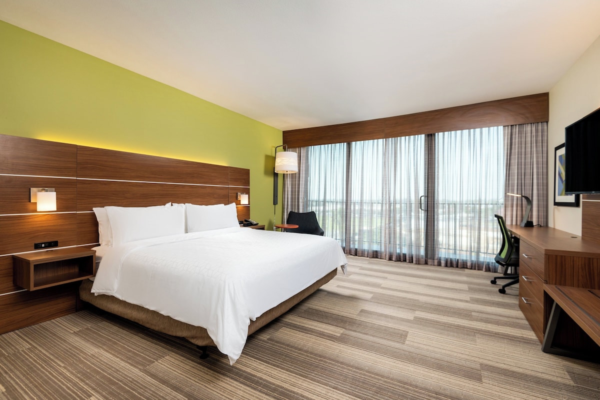 Habitación de hotel con una cama grande, un escritorio, una silla, un televisor y un balcón con cortinas.