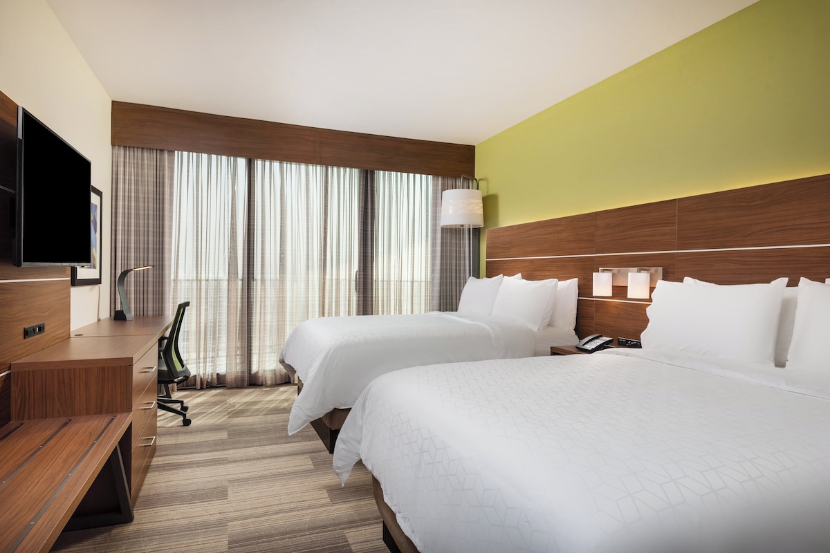 Habitación de hotel con dos camas, un escritorio, una silla, un televisor y un amplio ventanal con cortinas.