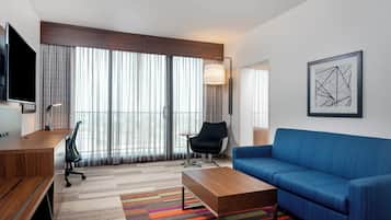A modern hotel room with a large window, a flat-screen TV, a sofa, a desk, and a colorful rug.