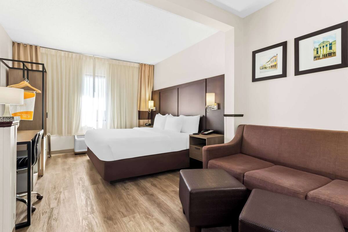 A hotel room with a large bed, a brown sofa, a desk with a chair, and two framed pictures on the wall.