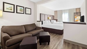 A hotel room with a brown sofa, a small ottoman, a bed with white linens, a headboard, a nightstand, and a TV on a stand. - Comfort Suites Lumberton North (Lumberton)