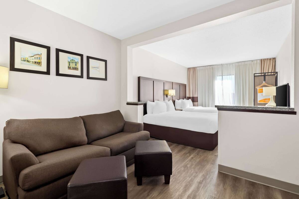 A hotel room with a brown sofa, a small ottoman, a bed with white linens, a headboard, a nightstand, and a TV on a stand.