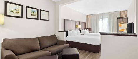 A hotel room with a brown sofa, a small ottoman, a bed with white linens, a headboard, a nightstand, and a TV on a stand.