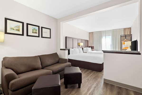 A hotel room with a brown sofa, a small ottoman, a bed with white linens, a headboard, a nightstand, and a TV on a stand.