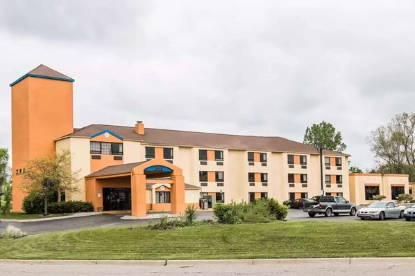 A multi-story hotel with a prominent orange tower, a central entrance with a blue awning, and a parking lot with several cars.
