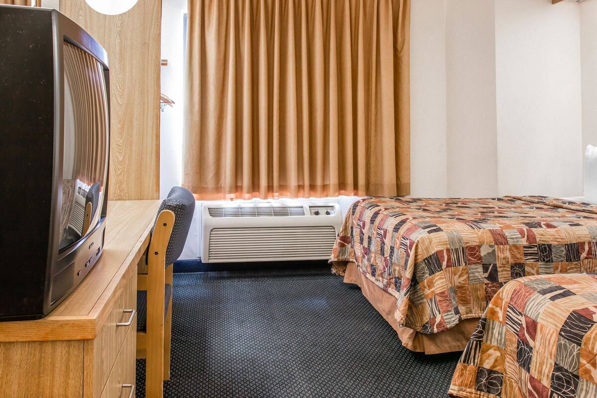Ein Hotelzimmer mit Fernseher auf einem Holztisch, einem Bett mit gemustertem Überwurf, einem Fenster mit Vorhängen und einer Wandklimaanlage.