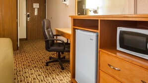 A hotel room with a wooden desk, a black office chair, a small refrigerator, a television, and a patterned carpet.