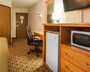 A hotel room with a wooden desk, a black office chair, a small refrigerator, a television, and a patterned carpet.