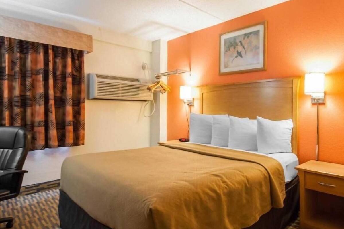 A hotel room with a bed, a chair, an air conditioning unit, a nightstand, and a wall-mounted lamp.