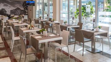 A modern restaurant with multiple tables and chairs, large windows, and indoor plants.