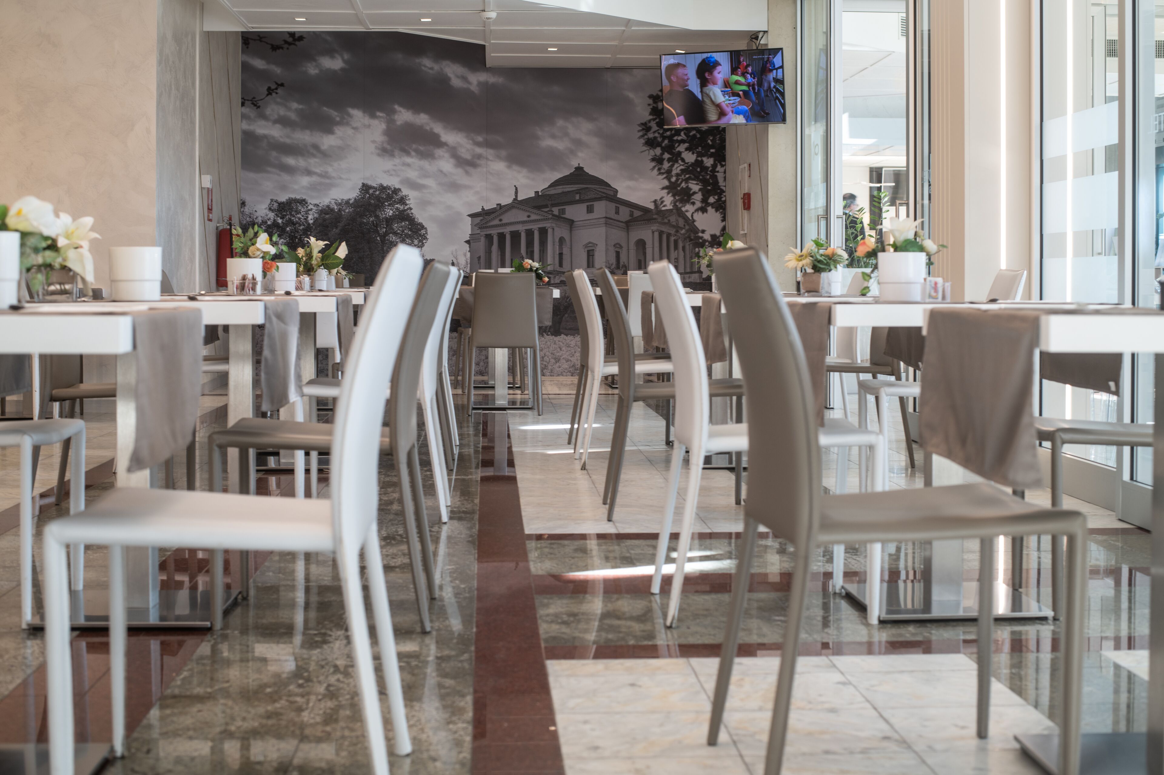 Un restaurante con una decoración moderna, que cuenta con una larga zona de comedor con sillas y mesas blancas, un mural de un edificio en la pared de fondo, y un televisor de pantalla plana montado sobre las mesas.