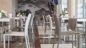 Un restaurante con una decoración moderna, que cuenta con una larga zona de comedor con sillas y mesas blancas, un mural de un edificio en la pared de fondo, y un televisor de pantalla plana montado sobre las mesas.