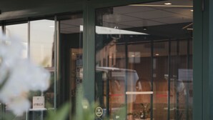 Une entrée d’immeuble avec une porte en verre, une enseigne au-dessus de la porte et le reflet d’un véhicule à l’intérieur.
