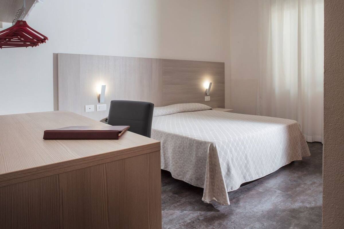 A hotel room with a wooden desk, a bed with a patterned bedspread, and a headboard with wall-mounted lights.