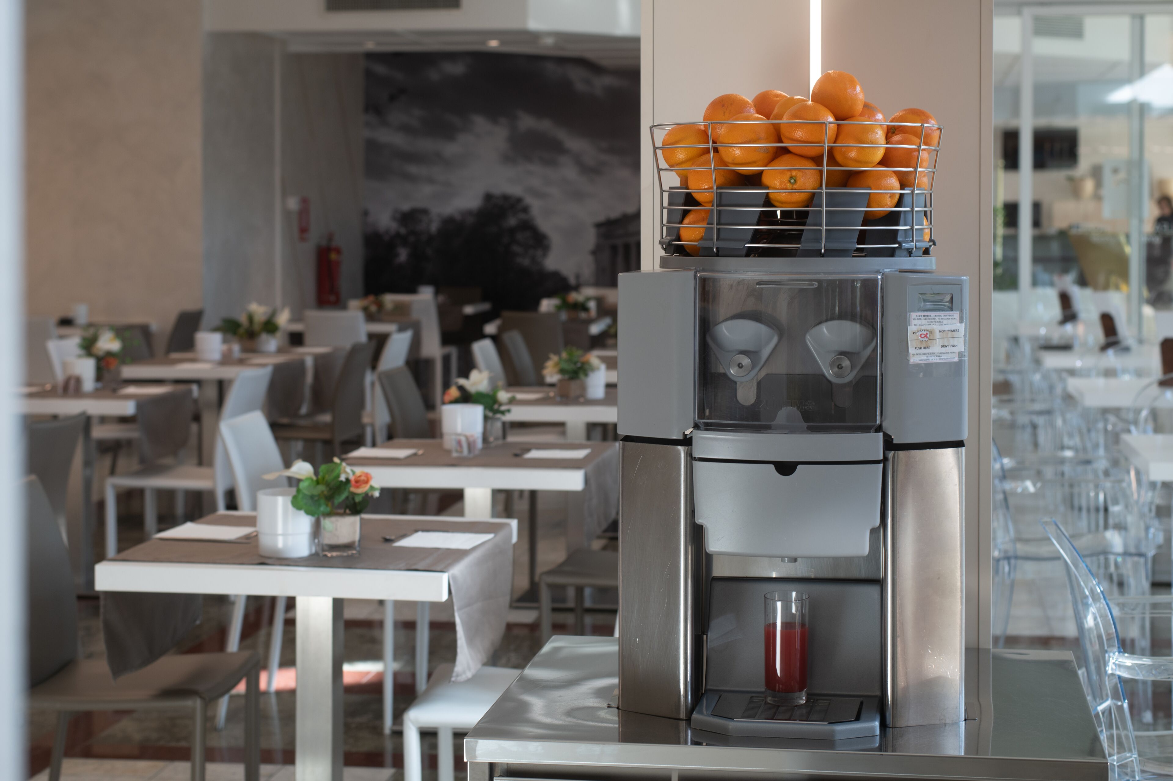 Una macchina da caffè con un cesto d'arance sopra, un bicchiere di bevanda rossa e una sala da pranzo moderna con tavoli e sedie.