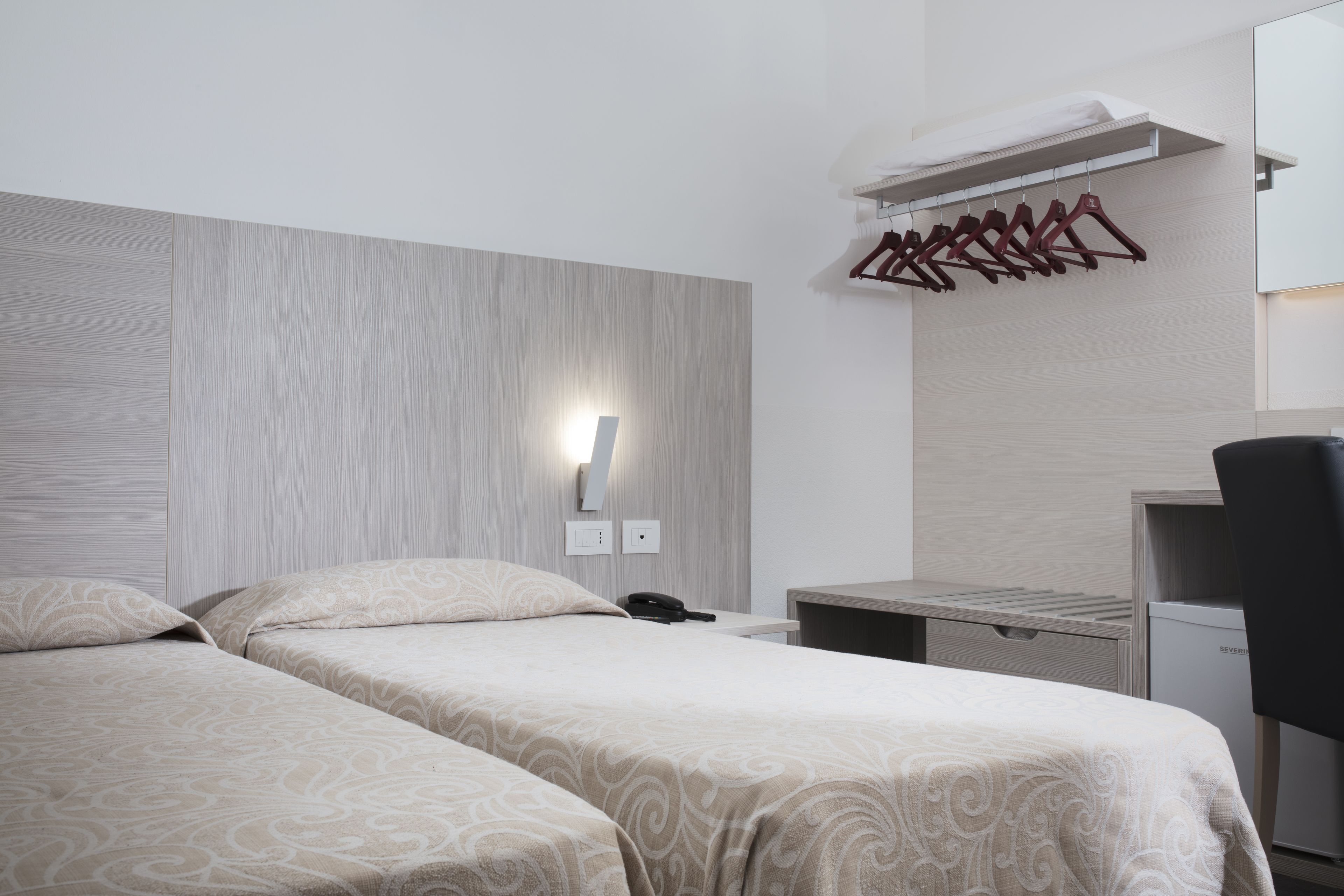 Une chambre d’hôtel avec deux lits, un bureau, une chaise, une armoire et une lampe fixée au mur.
