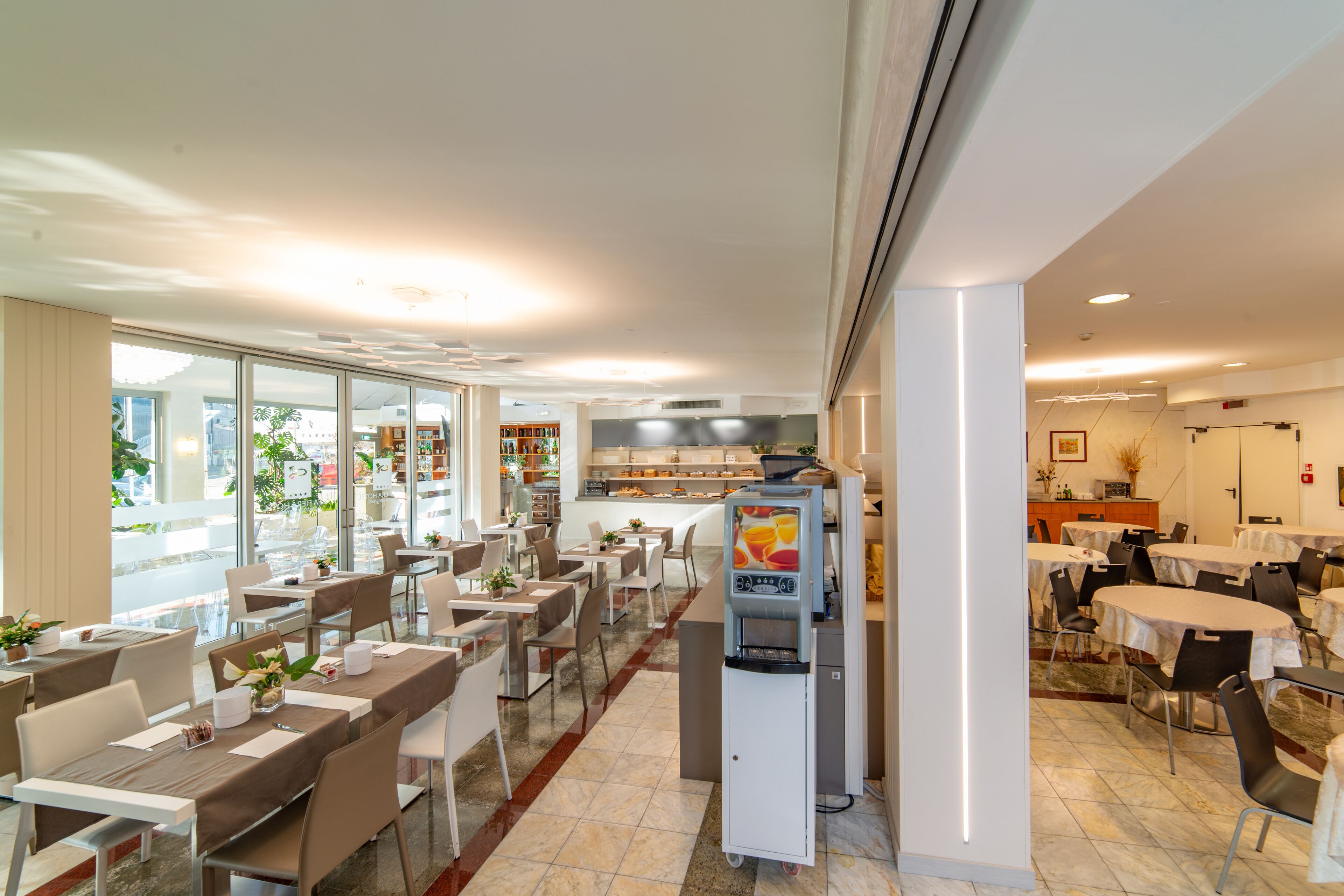 A modern dining area with tables and chairs, a juice dispenser, and a view of the street through large windows.