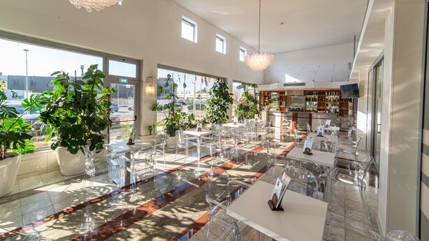 Un restaurant spacieux avec des tables et des chaises en verre transparent, de grandes fenêtres offrant une vue sur l’extérieur et une variété de plantes qui apportent une touche de verdure.