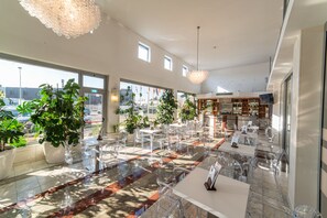 Een ruime restaurantruimte met transparante glazen tafels en stoelen, grote ramen met uitzicht naar buiten en diverse planten die groen toevoegen.