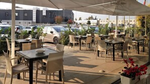 Un café en plein air avec des tables et des chaises, un espace abrité et une vue sur les voitures garées et les bâtiments.