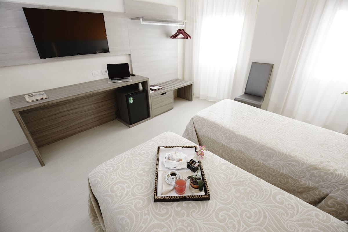 A hotel room with a bed, a desk with a laptop, a TV mounted on the wall, and a tray with breakfast items.