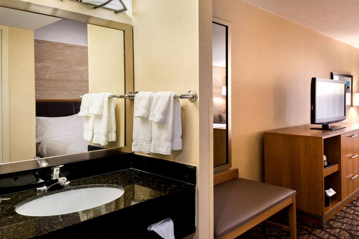 A hotel room with a large mirror, a sink, a wooden vanity, a television, a desk, and a chair.