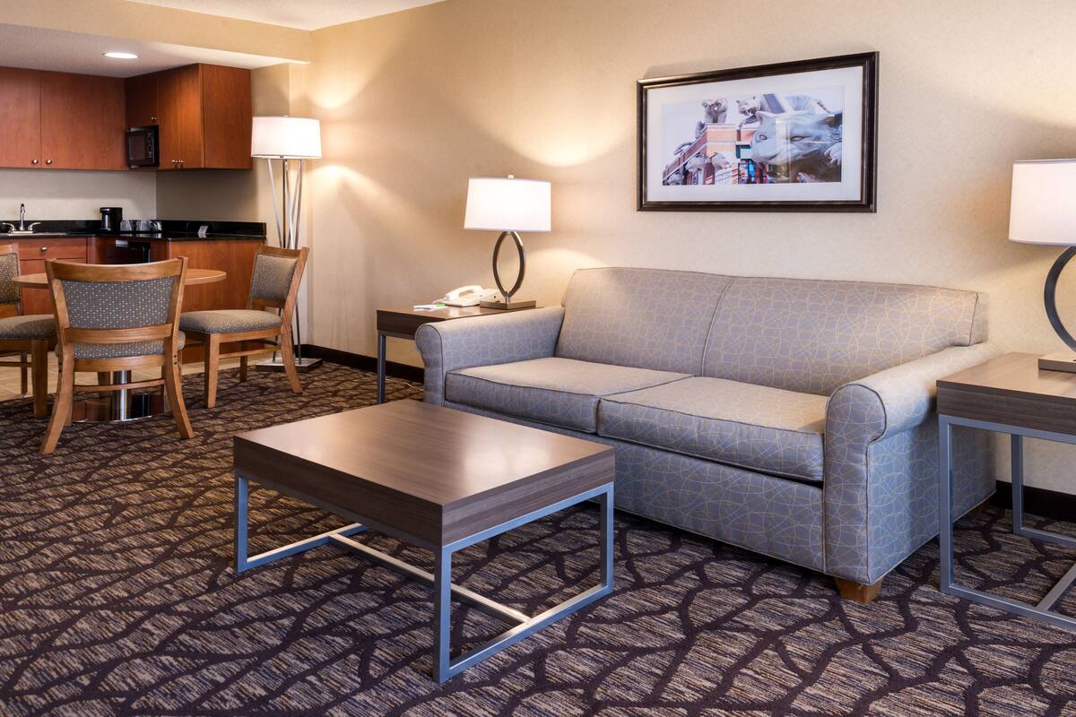 A hotel room with a sofa, two chairs, a coffee table, a lamp, a framed picture, and a kitchenette.