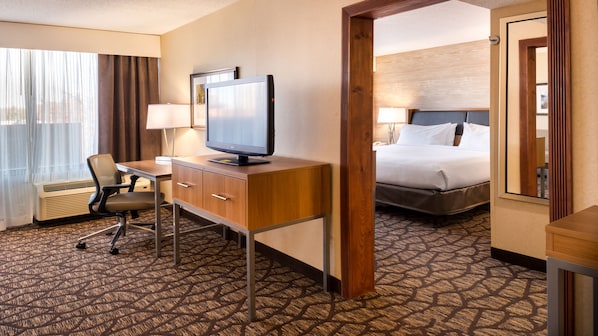 Quarto de hotel com escrivaninha, cadeira, televisão, cama e vista por uma porta.