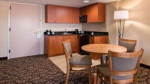 Quarto de hotel compacto com cozinha americana, área de refeições com mesa redonda e quatro cadeiras, e um abajur.