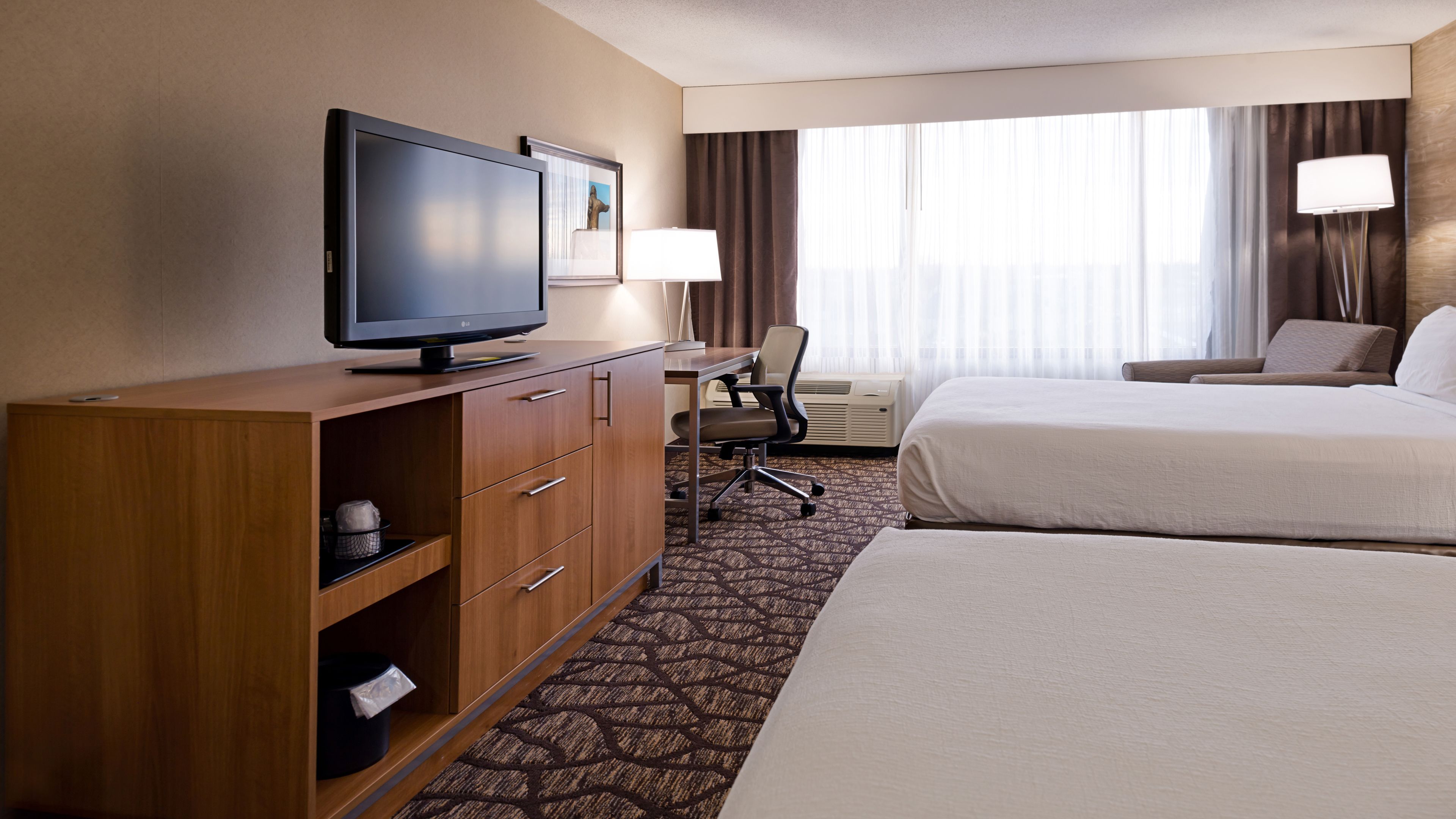 A hotel room with a flat-screen TV on a wooden cabinet, a desk with a chair, two beds, and a large window with curtains.