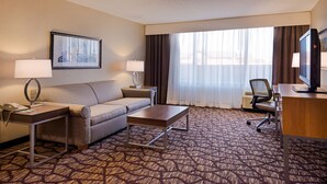 Quarto de hotel com sofá, mesa de centro, escrivaninha com computador, televisão e uma grande janela com cortinas.