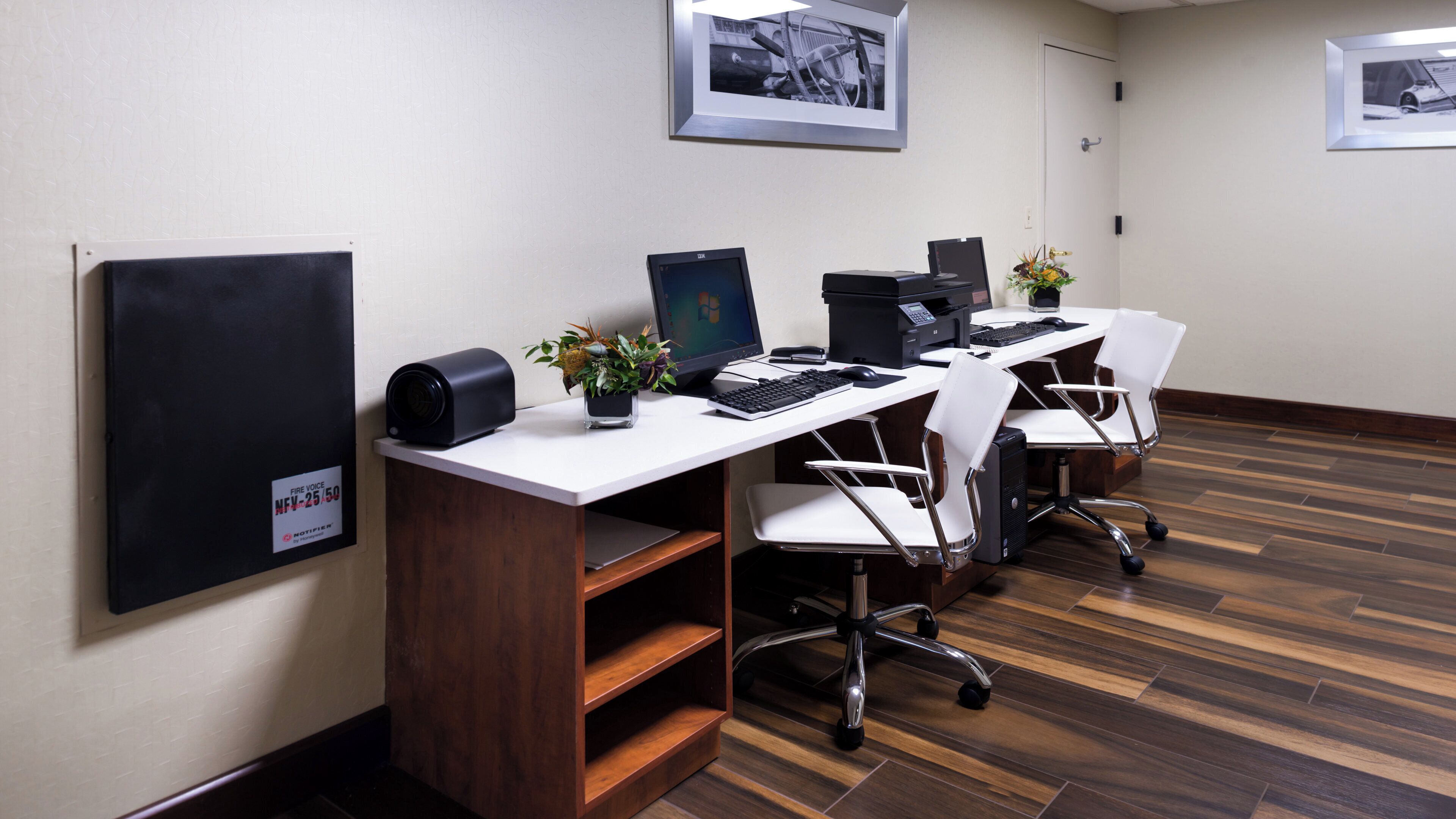 A conference room with three desks, each with a computer, keyboard, and mouse. There are two potted plants on the desks. The room has a wooden floor and white walls.