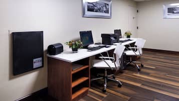 A conference room with three desks, each with a computer, keyboard, and mouse. There are two potted plants on the desks. The room has a wooden floor and white walls.
