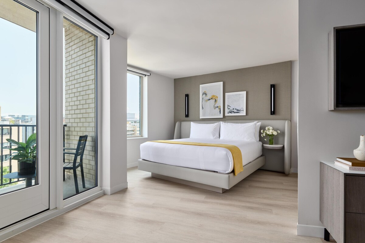 A modern bedroom with a large bed, a bedside table with a vase of flowers, and a view of the city through a sliding glass door.