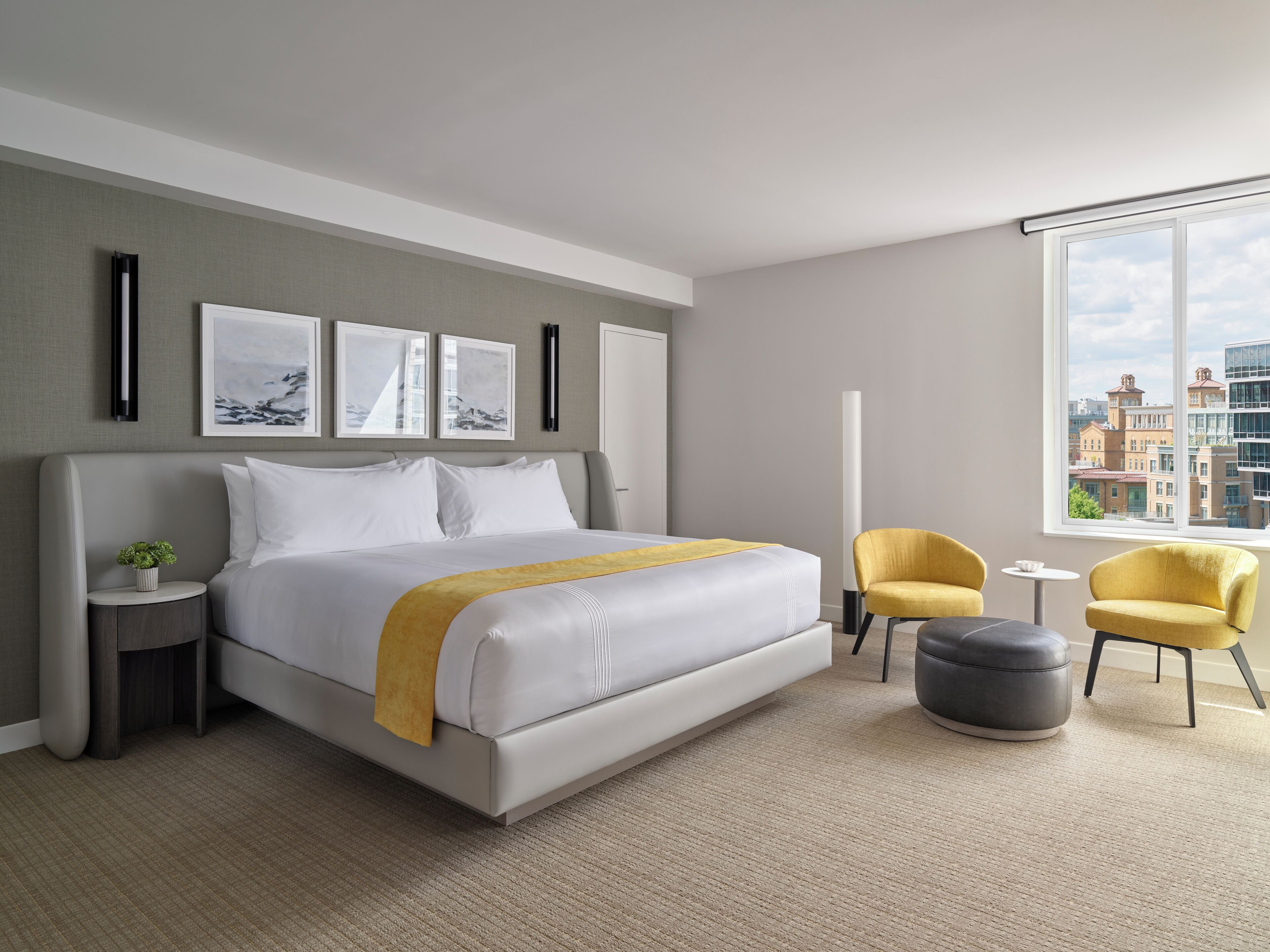 Una habitación de hotel moderna con una cama grande, una mesita auxiliar con una planta, un sillón amarillo, un reposapiés gris y vistas a la ciudad a través de un ventanal amplio.