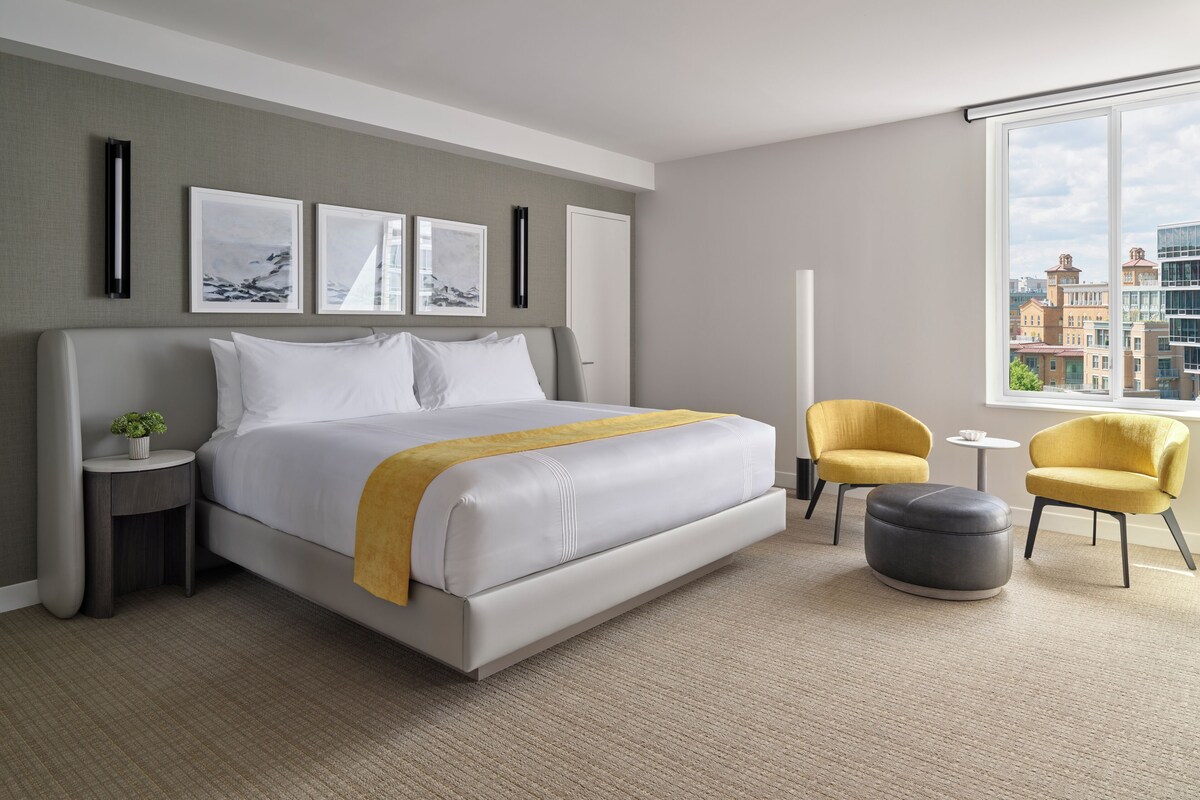 A modern hotel room with a large bed, a small side table with a plant, a yellow armchair, a grey ottoman, and a city view through a large window.
