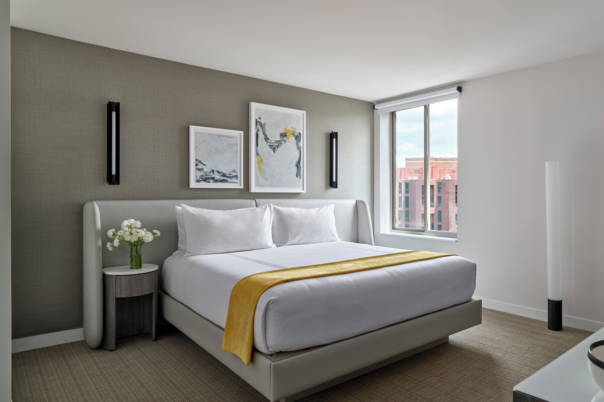 A modern hotel room with a large bed, a bedside table with a vase of flowers, and artwork on the wall.