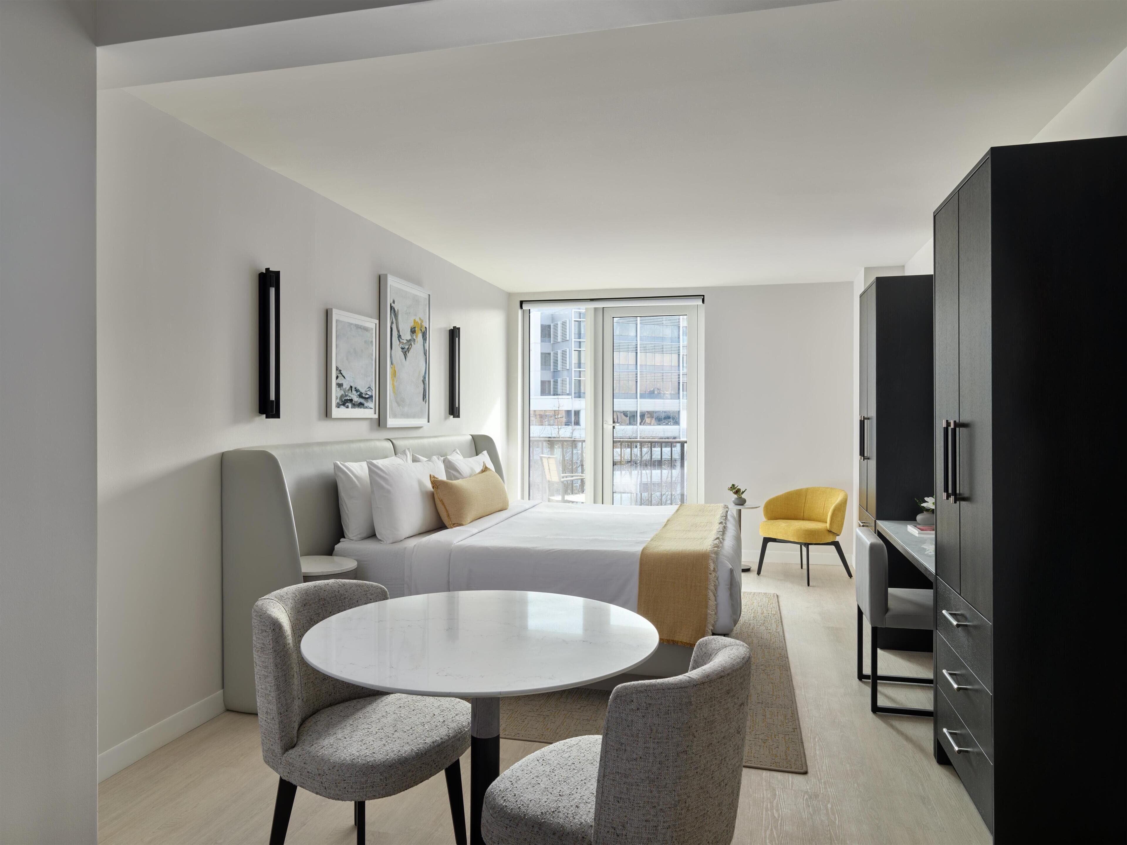 A modern hotel room with a bed, a round dining table, two armchairs, a sofa, a yellow chair, and a wardrobe.