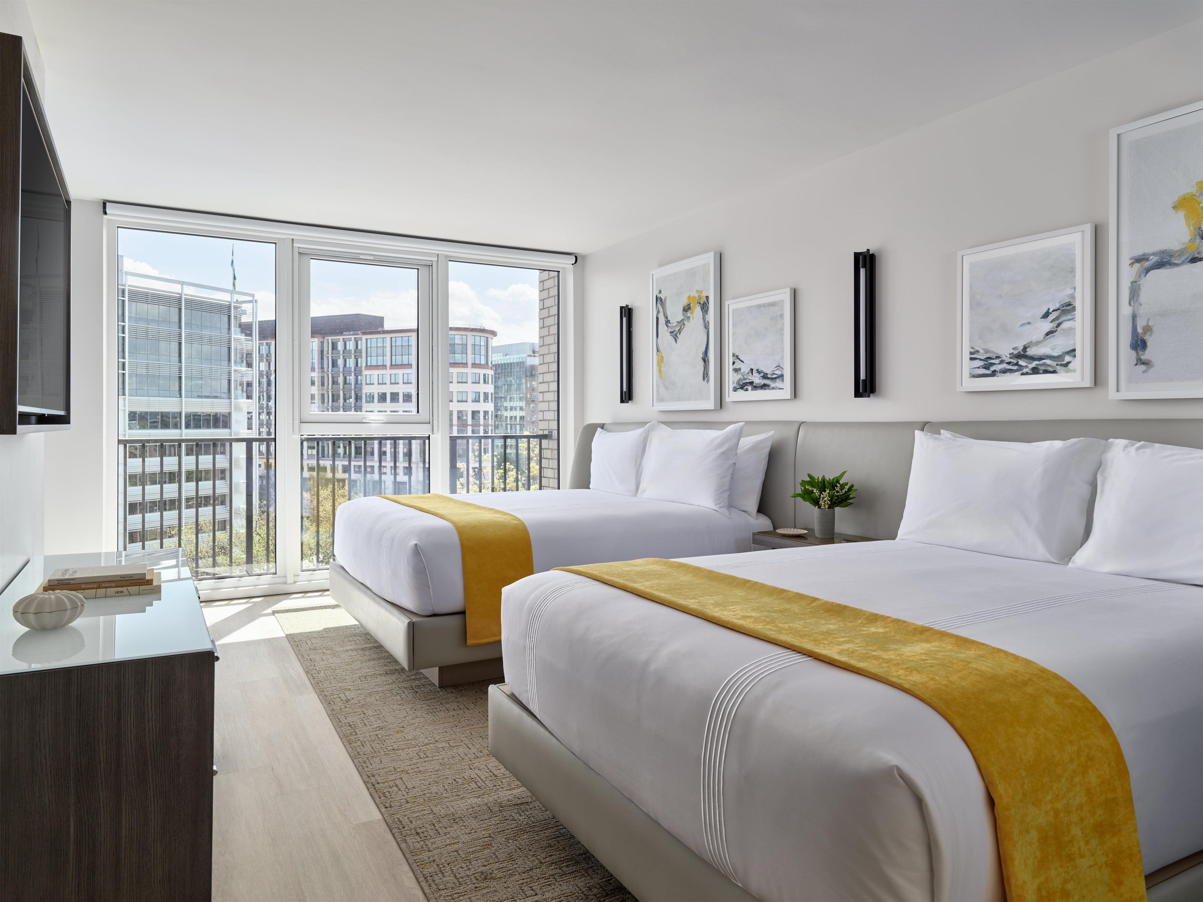 A hotel room with two beds, a desk, a potted plant, and artwork on the walls.