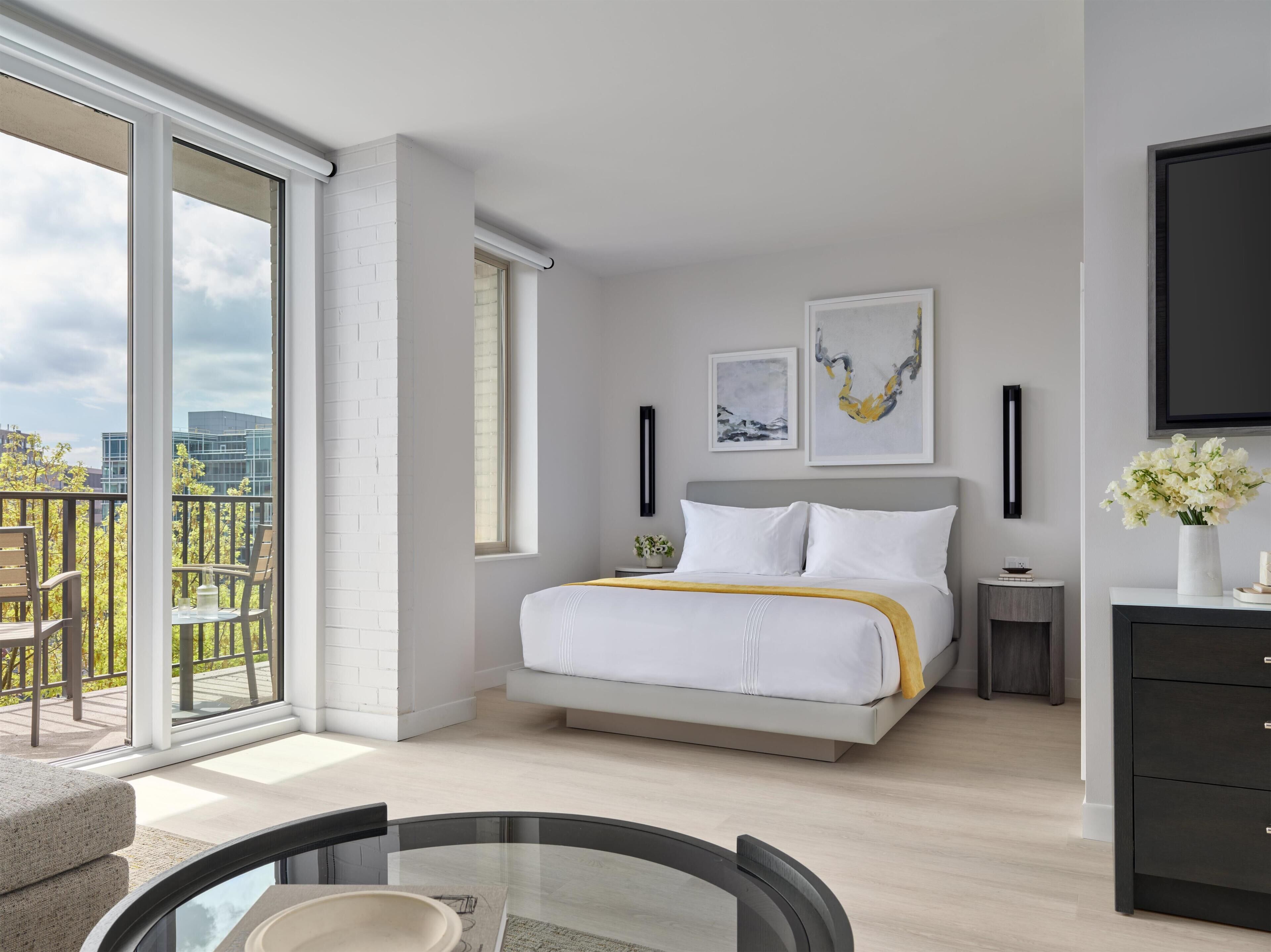 A modern bedroom with a large bed, a bedside table, a TV, and a balcony with outdoor furniture.