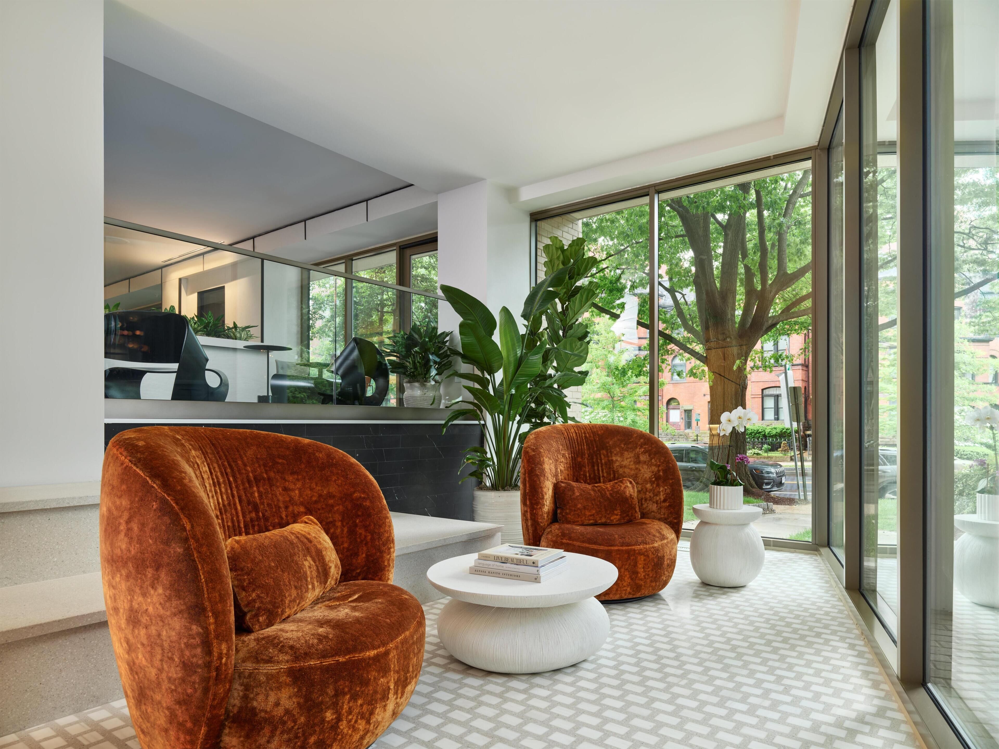 A modern living room with two large velvet armchairs, a white coffee table, and a large window with a view of trees and buildings.