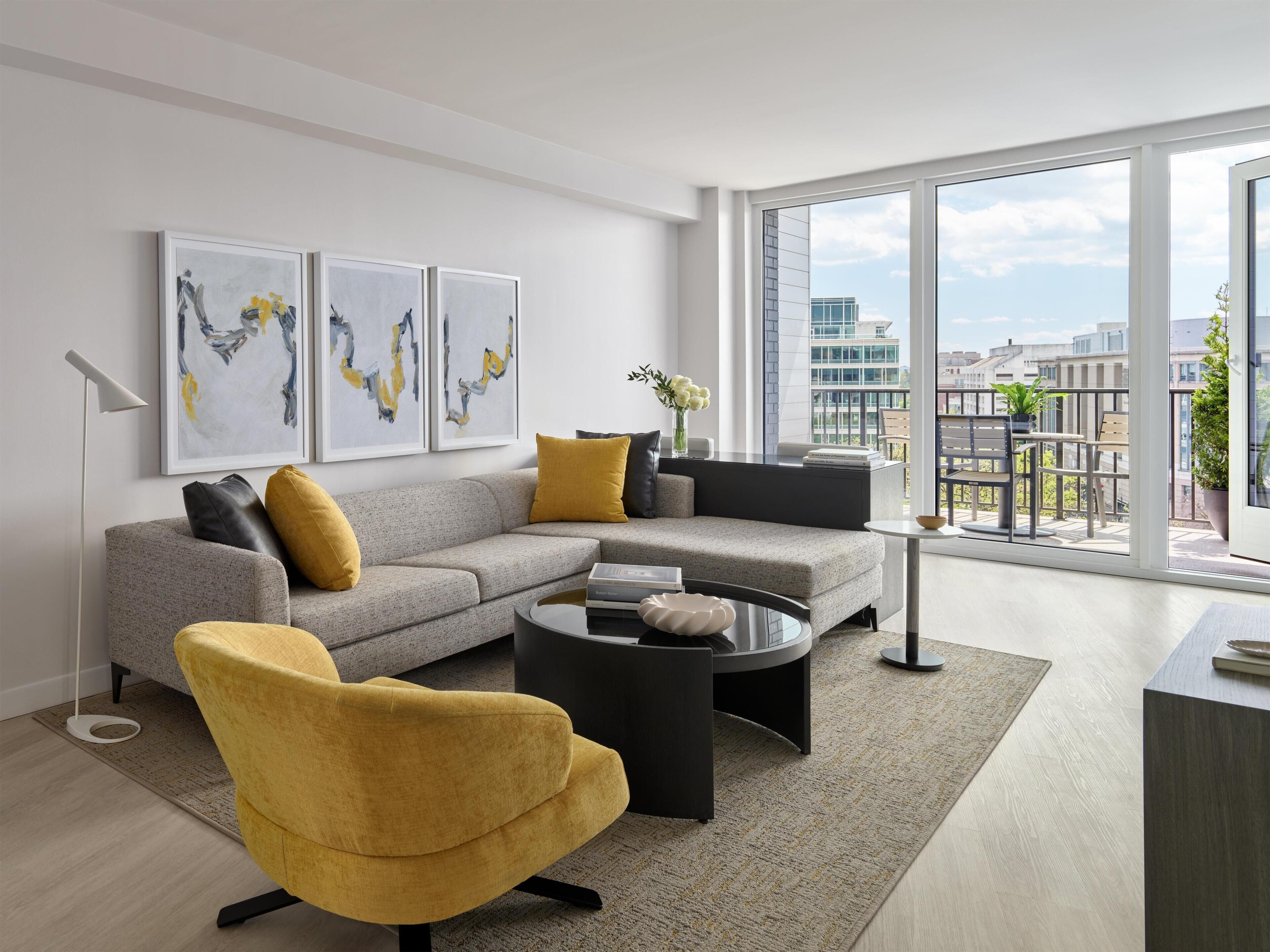 A modern living room with a grey sofa, yellow armchair, and a coffee table. There are abstract paintings on the wall and a view of the city through large windows.