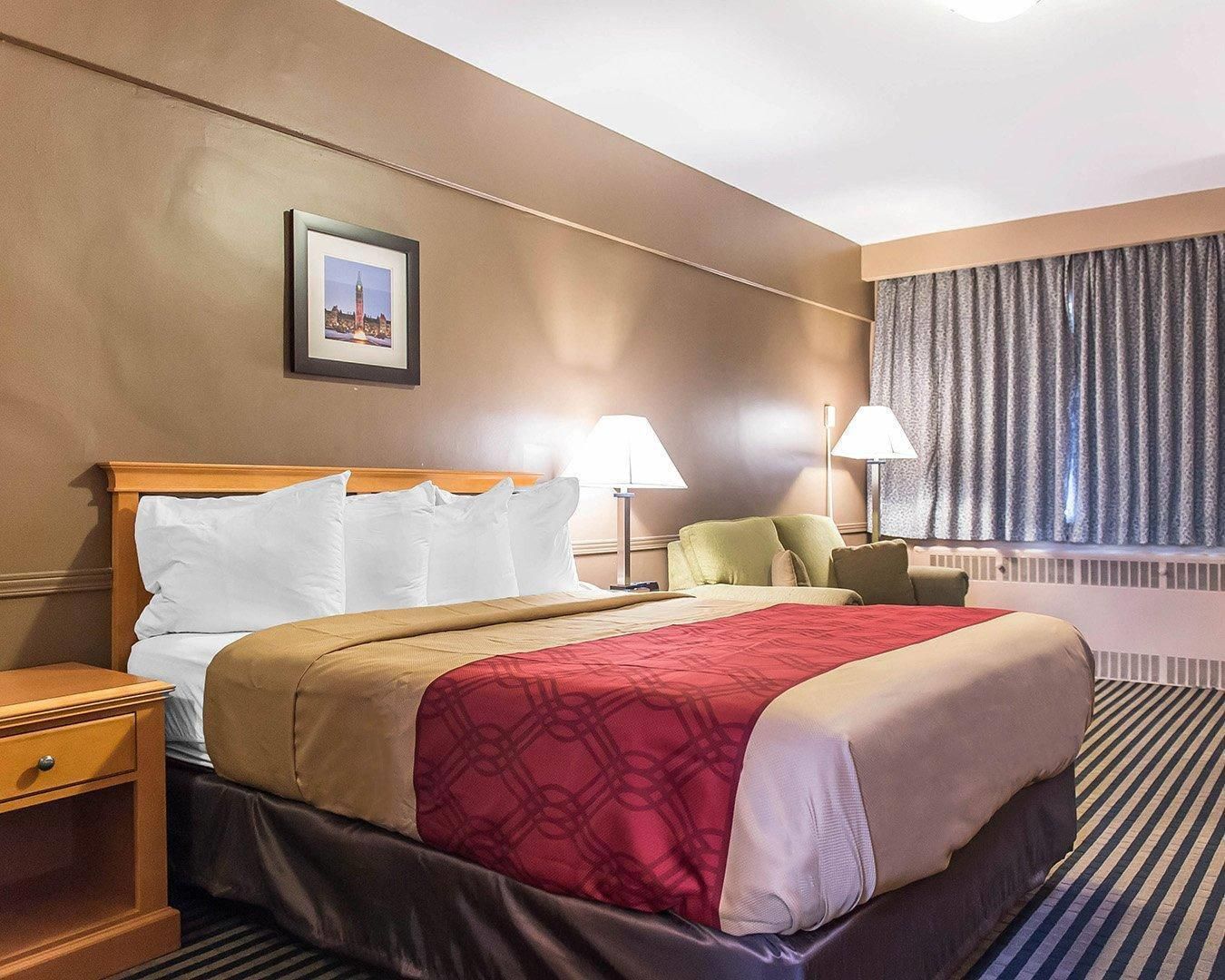 Une chambre d’hôtel avec un lit, des tables de chevet, des lampes, une fenêtre avec des rideaux et un tableau encadré au mur.