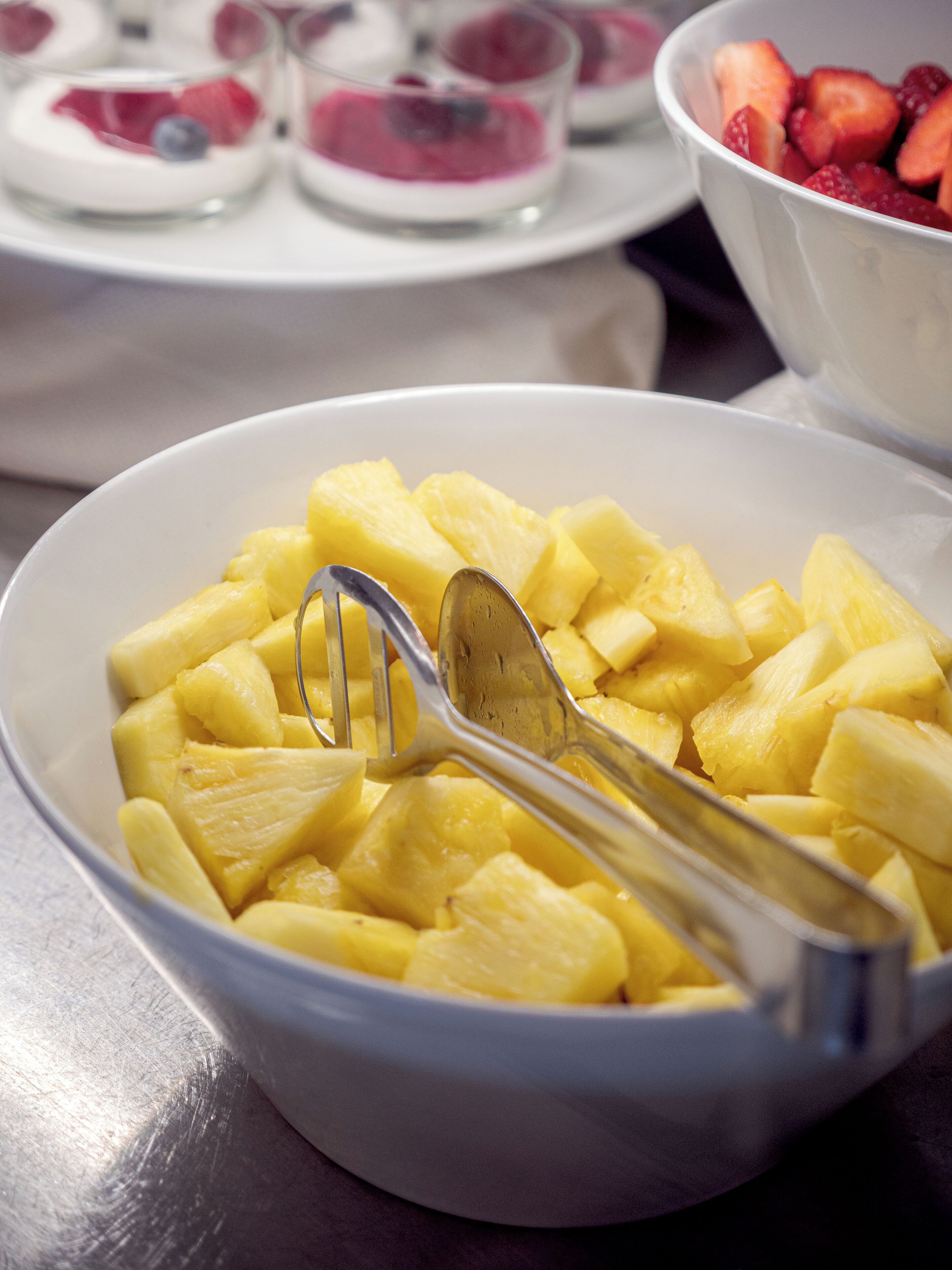 A bowl of diced pineapple with a spoon, and a bowl of sliced strawberries in the background.