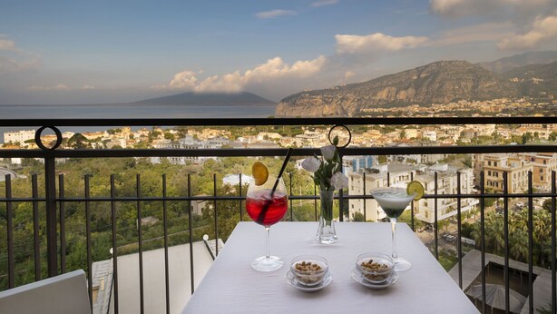 Un balcón con una mesa dispuesta para dos personas, con vistas a una ciudad costera y montañas al fondo.