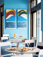 A dining area with a table set for breakfast, featuring plates of food, glasses, and a bottle of orange juice. There are two large paintings on the wall depicting a blue sky and ocean scene.