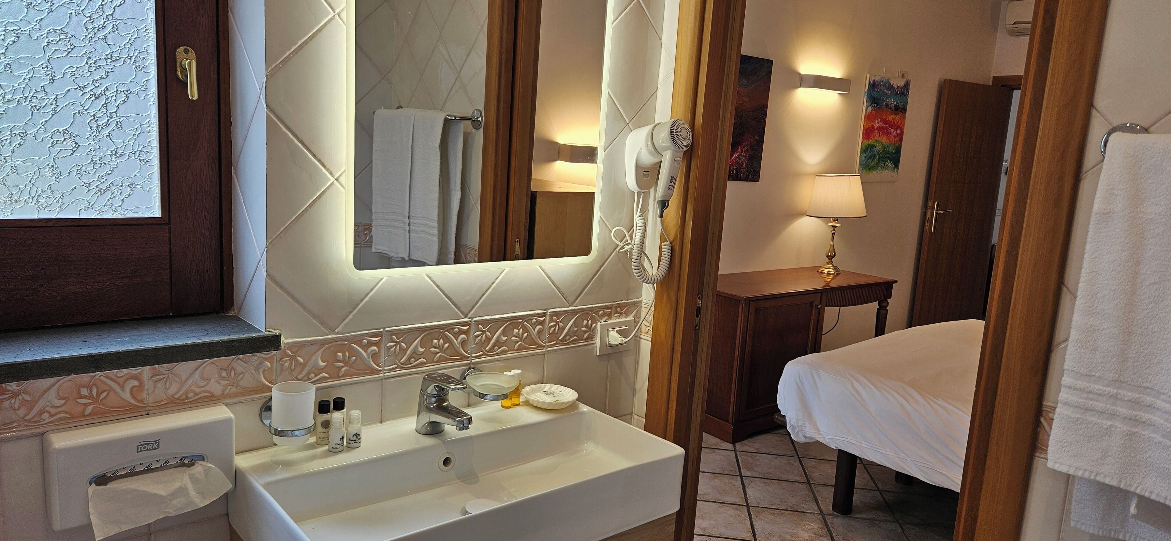A bathroom with a sink, mirror, hairdryer, and a bed with a towel.