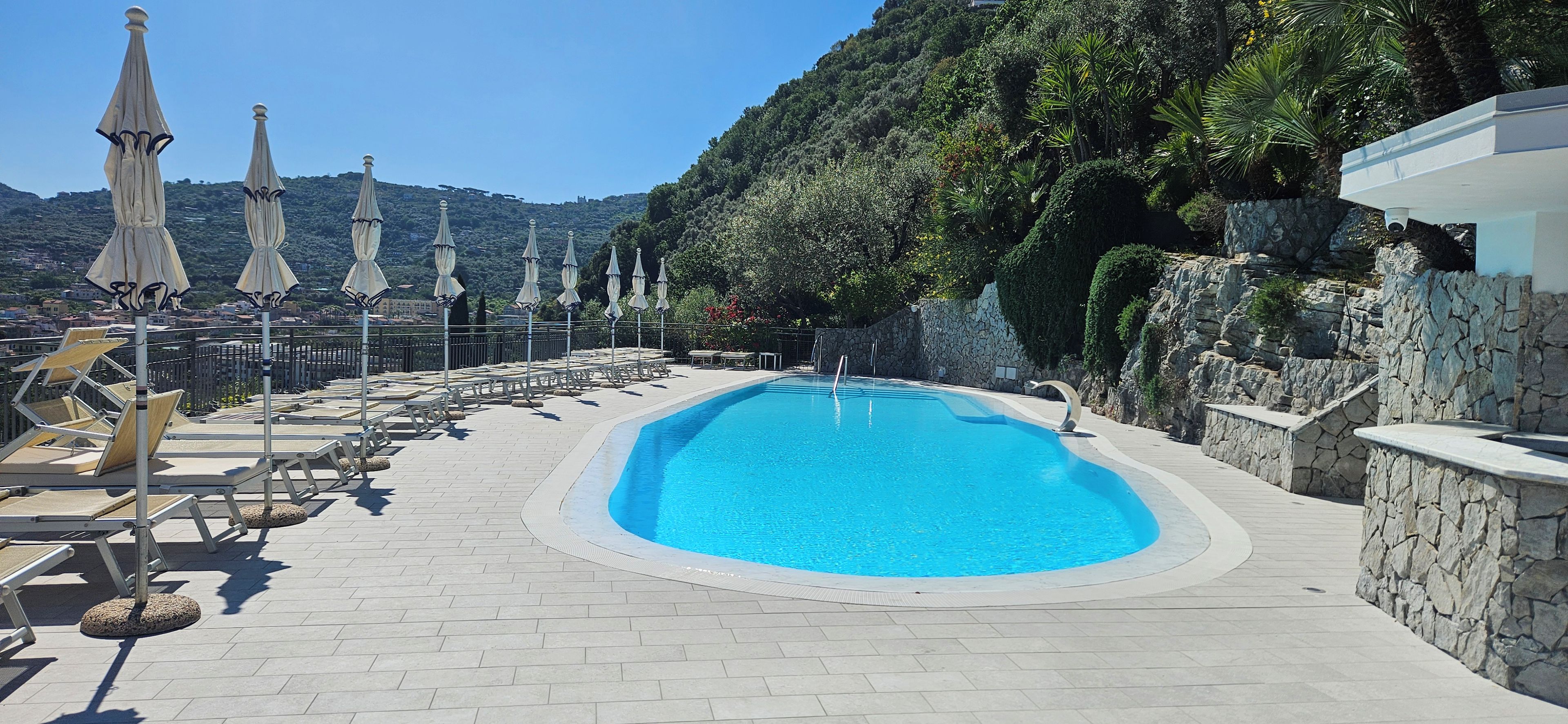 Una piscina con sillas de descanso y sombrillas, rodeada de muros de piedra y vegetación.